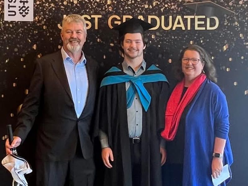 Tobias in graduation robes between two people in front of a UTS Just Graduated backdrop